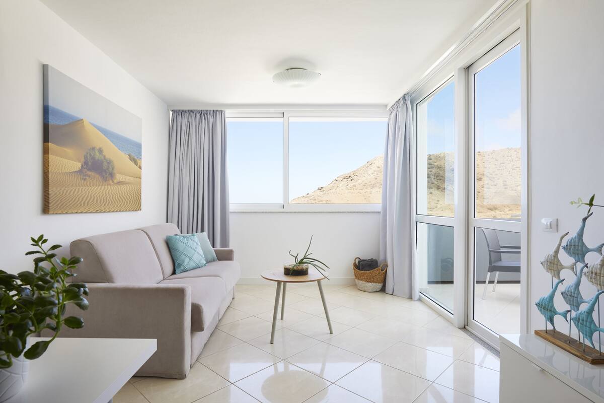 A modern living room with a beige sofa, a small round table with a plant, and a large window offering a view of the desert.