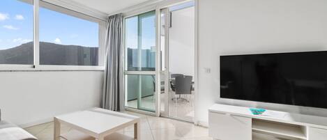 A modern living room with a large flat-screen TV, a white sofa, a small table, and a sliding glass door leading to a balcony with a city view.