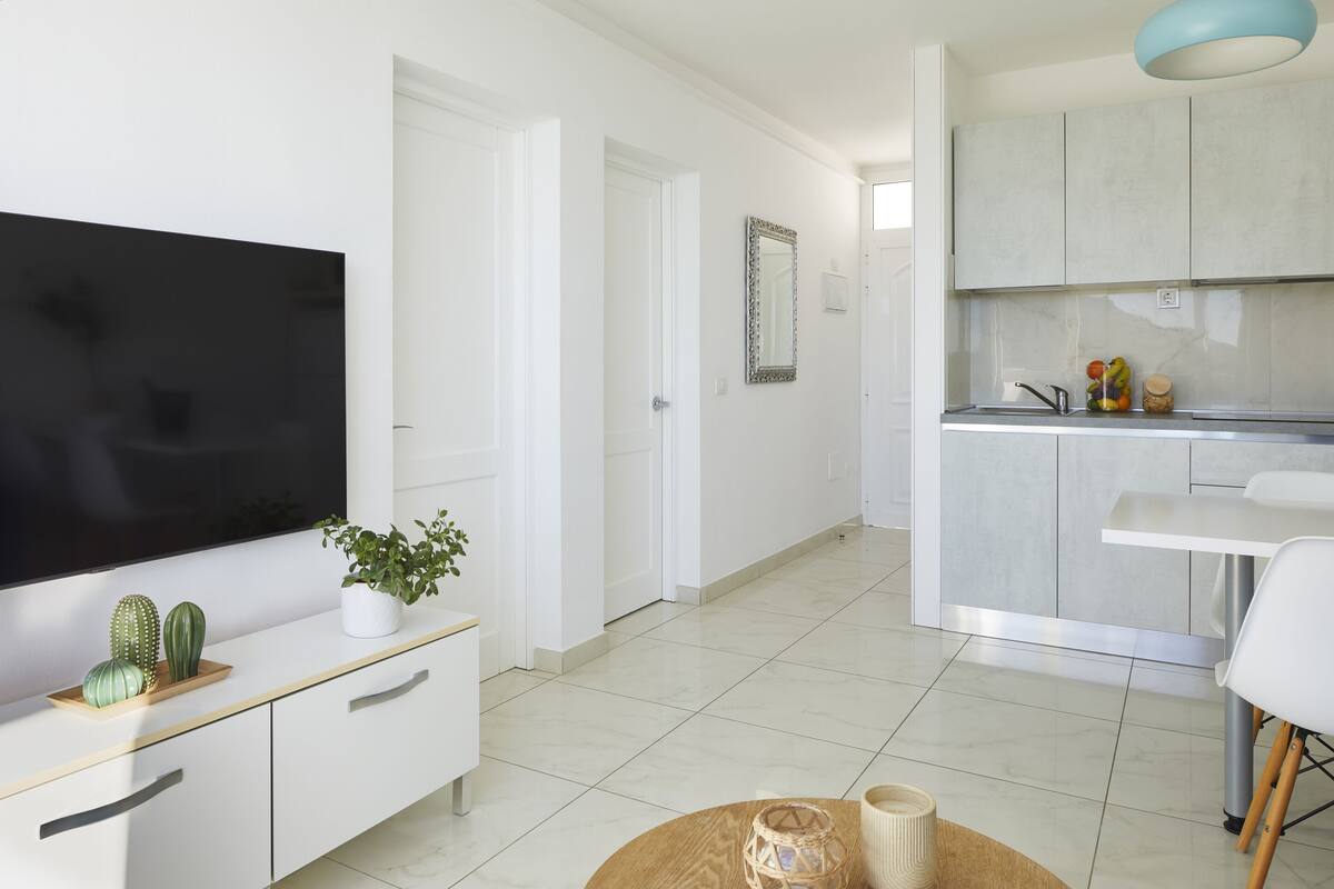 A modern kitchen with white cabinets, a dining area with a white table and chairs, a flat-screen TV on a white console, and a potted plant.