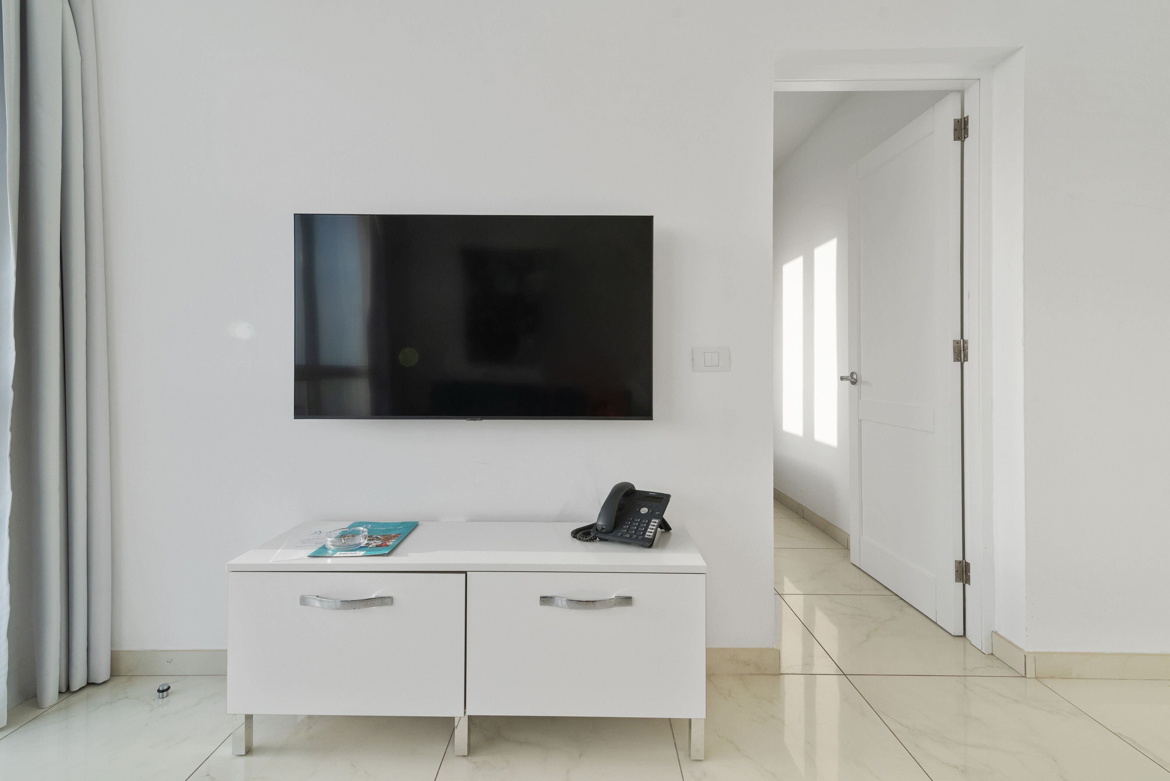 A modern hotel room with a flat-screen TV mounted on the wall, a white console table with a telephone and a magazine, and a door leading to another room.