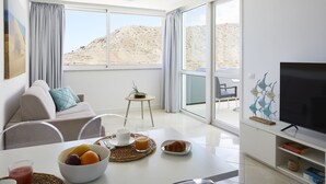 A modern hotel room with a dining area, a sofa, a TV, and a balcony with a desert view.