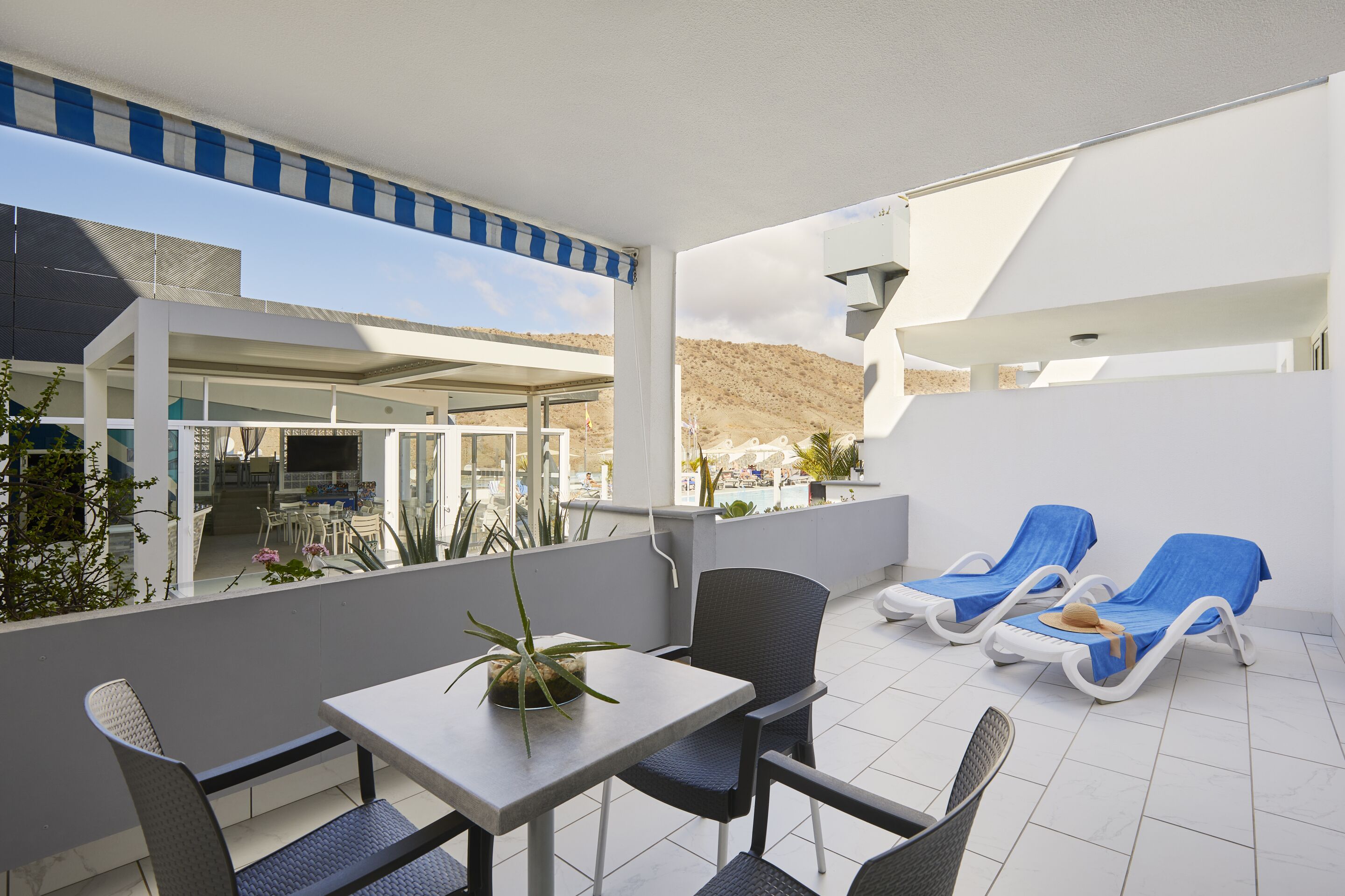 A terrace with a table and chairs, two sun loungers, and a view of a pool and mountains.