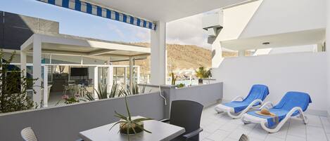 A terrace with a table and chairs, two sun loungers, and a view of a pool and mountains.