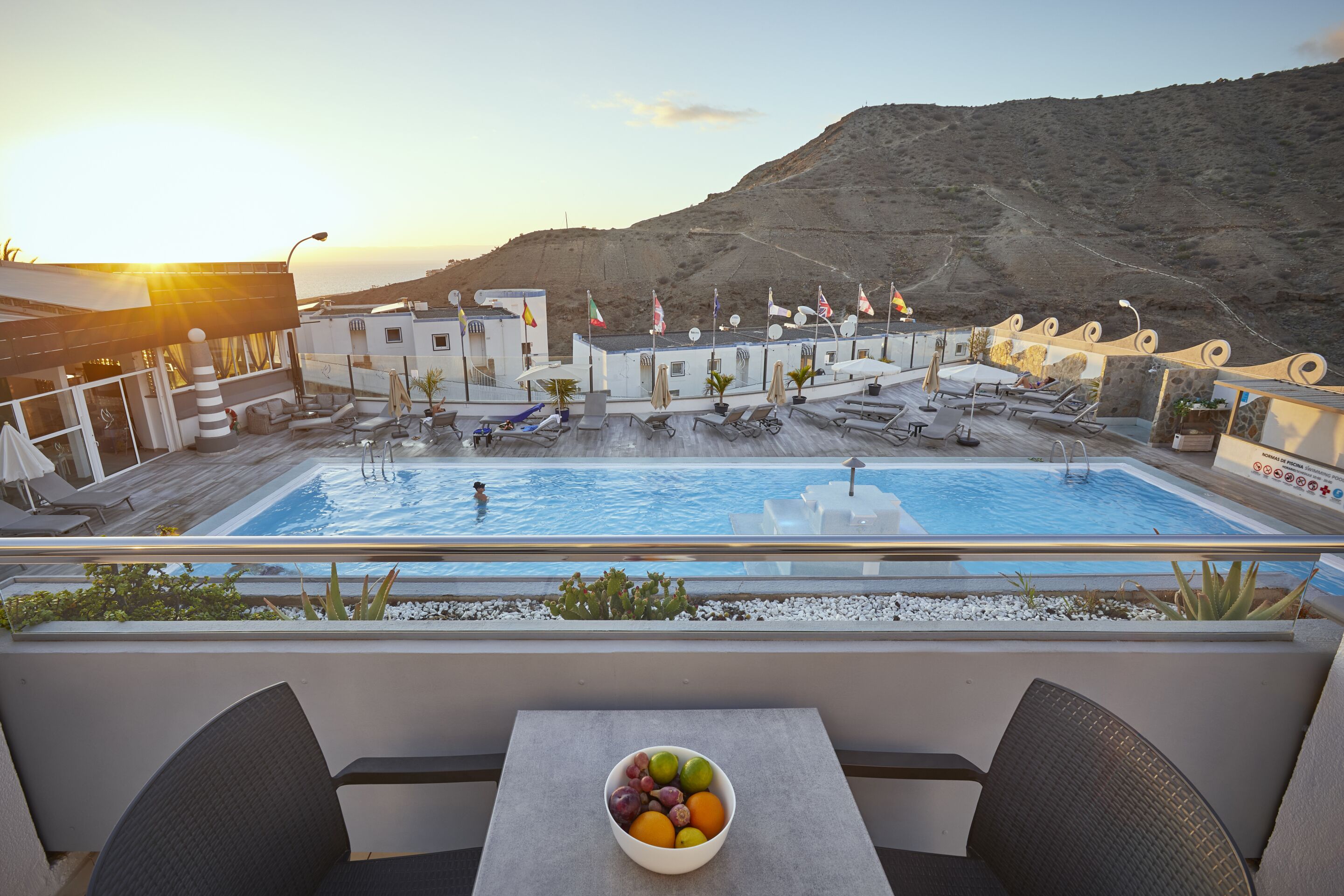 Un espace piscine aménagé sur le toit, avec des chaises longues, une table sur laquelle est posé un panier de fruits et des drapeaux.