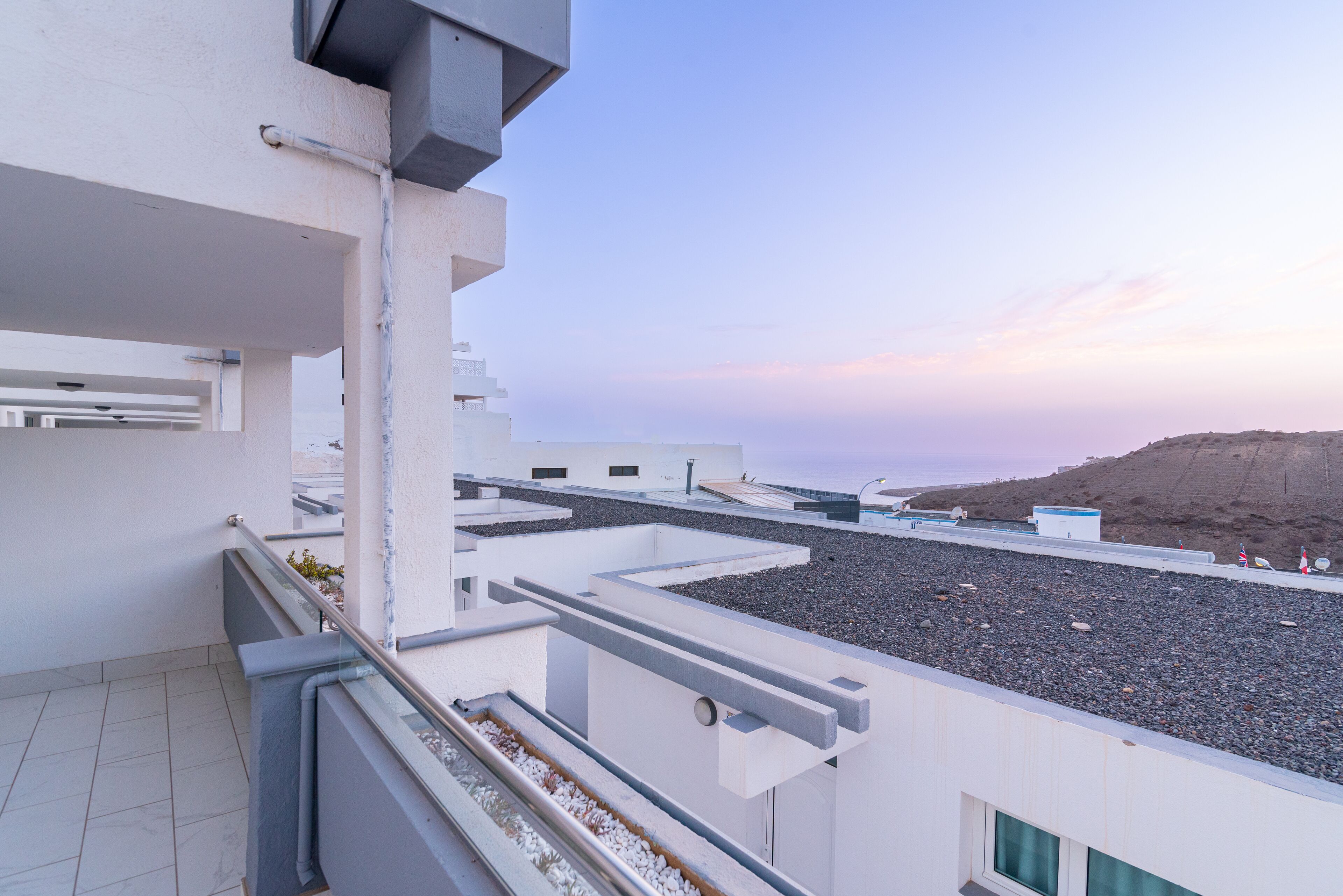 A modern building with a rooftop terrace overlooking a pebble beach and a distant hill.