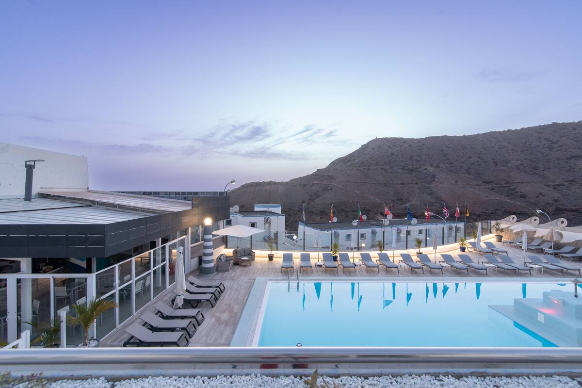 A rooftop pool area with lounge chairs and umbrellas, overlooking a mountainous landscape.