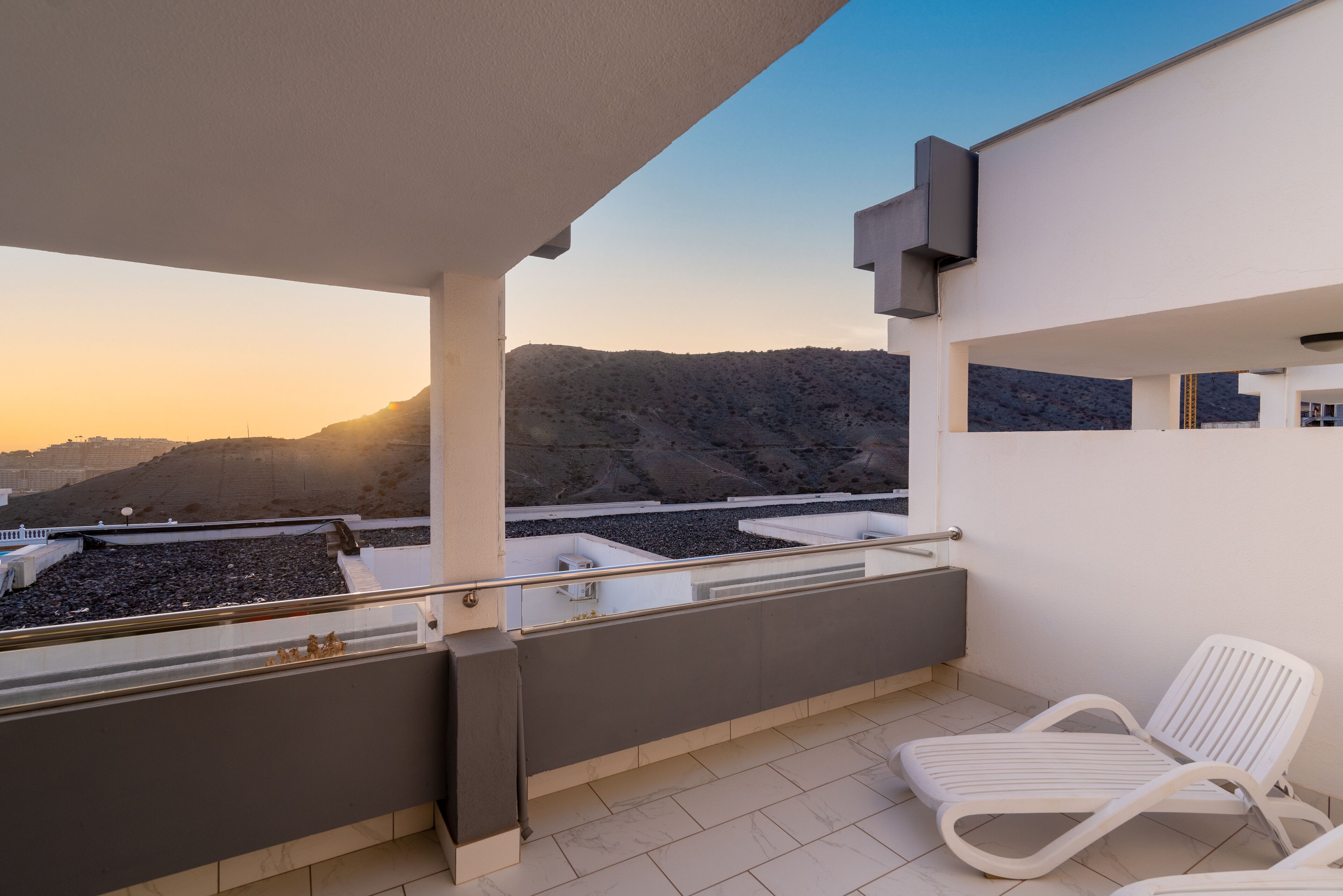 Un balcón con una silla blanca, vistas de un paisaje montañoso y un edificio moderno de techo plano.