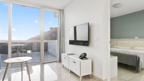A modern hotel room with a large sliding glass door leading to a balcony, a flat-screen TV mounted on the wall, a small white bedside table, and a bed with a grey bedspread.