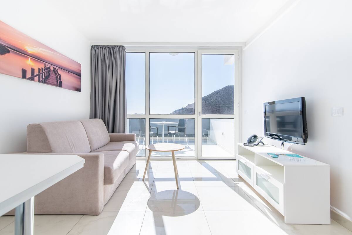 A modern living room with a sofa, a coffee table, a TV, and a balcony with a view of the mountains.