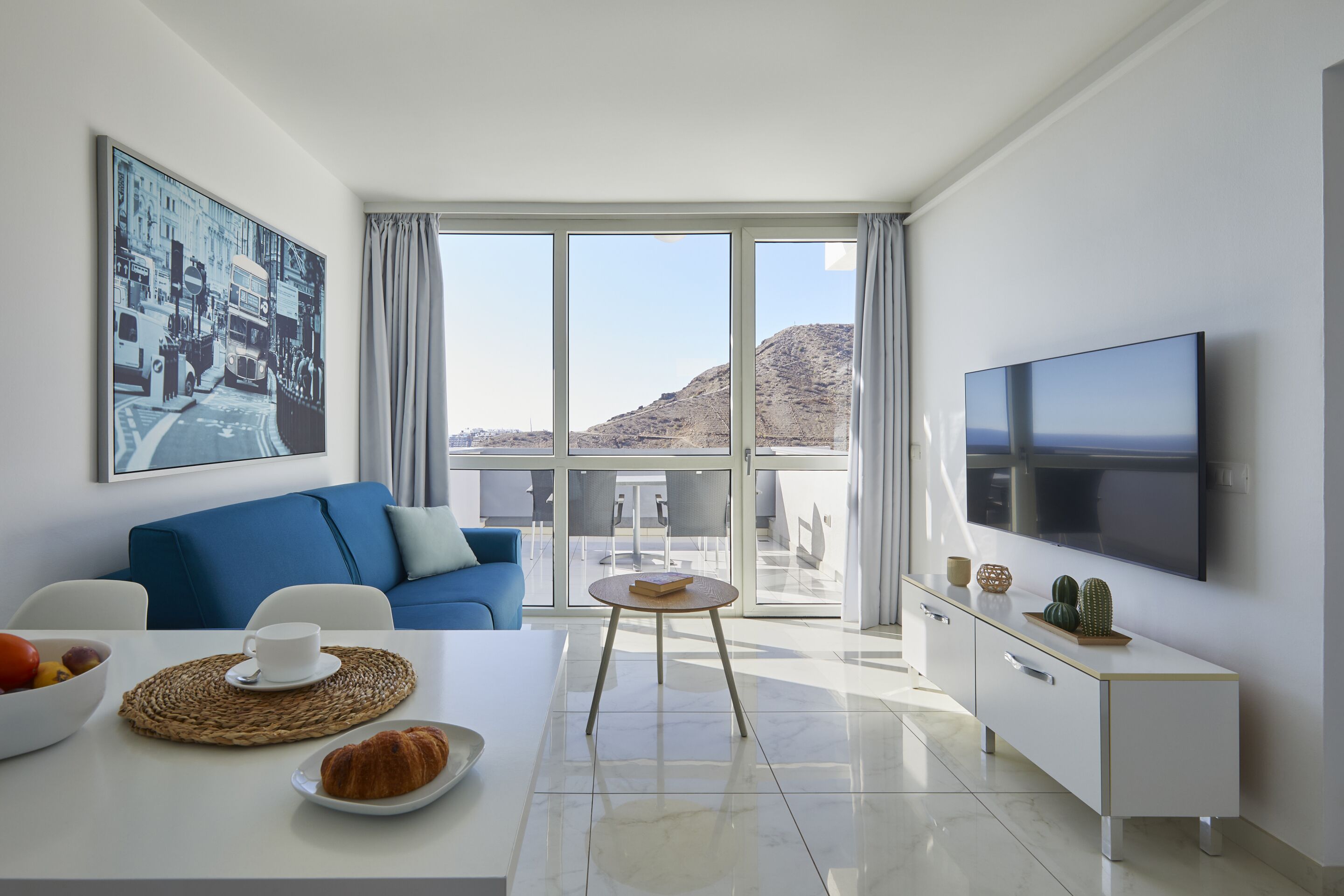 A modern living room with a blue sofa, a dining table with a croissant and fruit bowl, a TV, and a balcony with a mountain view.