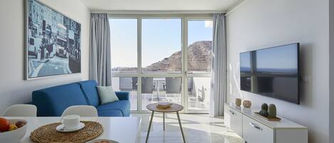 A modern living room with a blue sofa, a dining table with a croissant and fruit bowl, a TV, and a balcony with a mountain view.