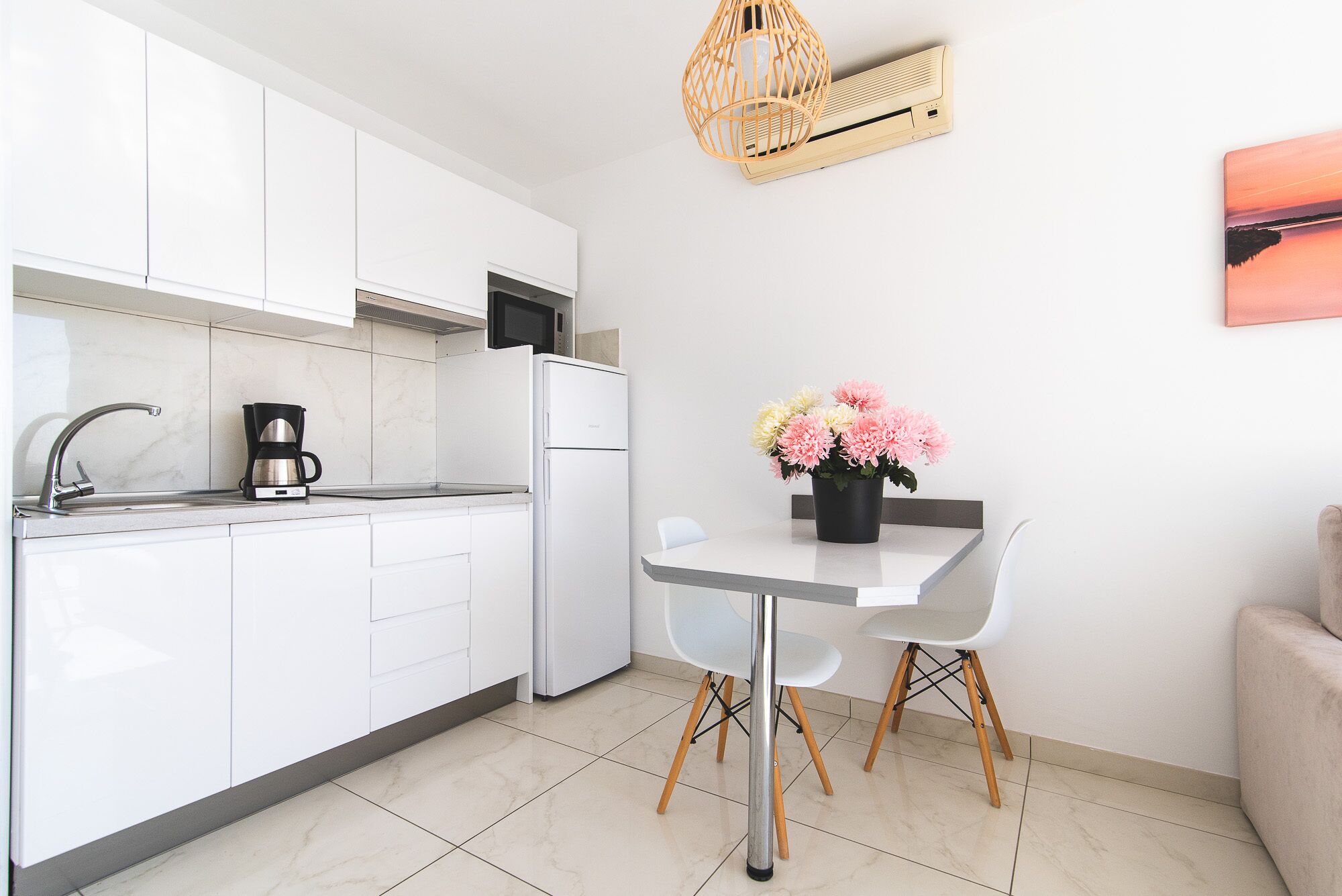 A modern kitchen with white cabinets, a built-in microwave, and a coffee maker. There is a dining table with two chairs and a vase of flowers.