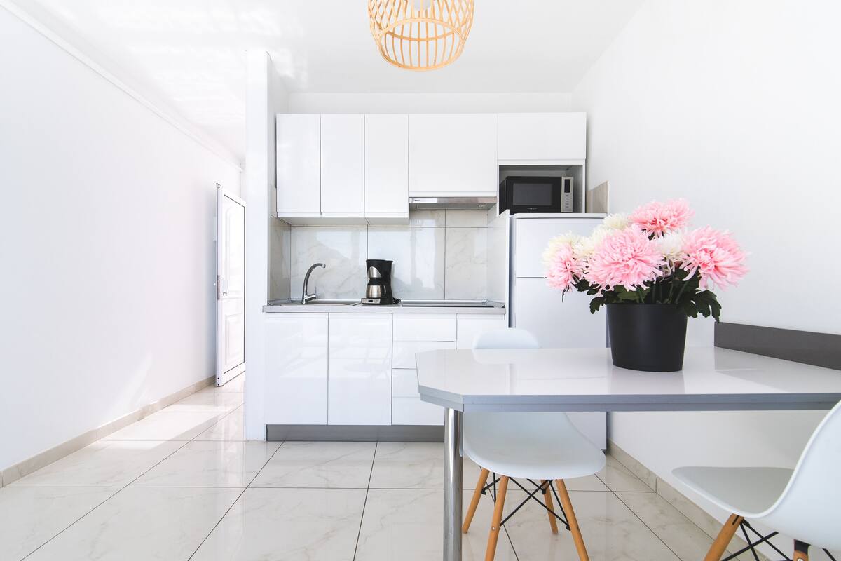 A modern kitchen with white cabinets, a stainless steel refrigerator, and a black coffee maker. There is a table with two chairs and a potted plant with pink flowers.