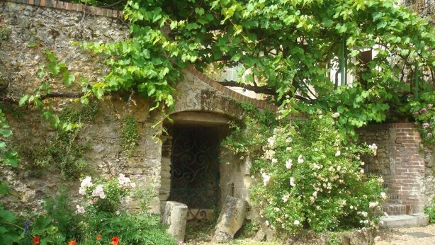 A stone archway with a vine-covered entrance, surrounded by flowering plants and a gravel path.