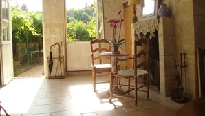 A room with a wooden chair, a small table, a window with a view of greenery, and a fireplace with a vase on top.