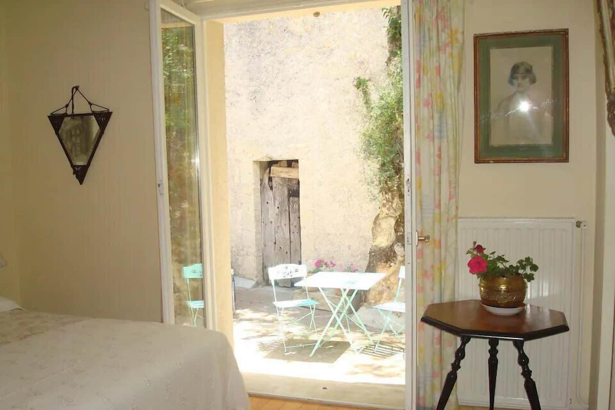 Un dormitorio con cama, una mesita de noche con un florero, y vista a un patio exterior con sillas y una mesa.