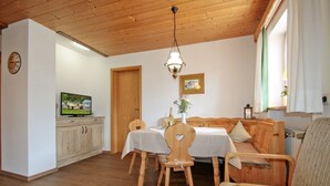 Ein gemütliches Wohnzimmer mit Holzbalkendecke, einem Esstisch mit Stühlen, einer Couch, einem Fernseher und einem Fenster mit Vorhängen.