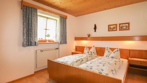 Ein Schlafzimmer mit einem hölzernen Bett, einem Nachttisch, einer Kommode, einem Fenster mit Vorhängen, einem Heizkörper und zwei gerahmten Bildern an der Wand.