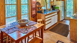 Cocina de una cabaña de madera con una mesa de comedor, un refrigerador y una ventana con vistas a un bosque.