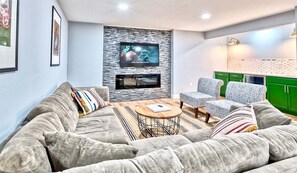 Una sala de estar moderna con un sofá gris, sillones estampados, una mesa de centro y un televisor de pantalla plana montado en una pared de piedra.