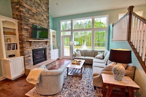 A living room with a stone fireplace, a television, a sofa set, and a large window with a view of trees.
