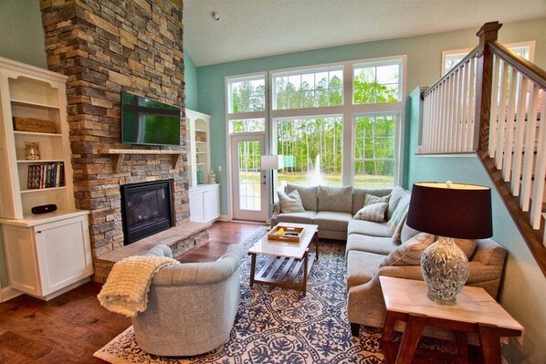 A living room with a stone fireplace, a television, a sofa set, and a large window with a view of trees.