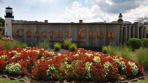 Une enseigne en bois sur laquelle est inscrit « COASTAL CLUB », entourée d’un jardin agrémenté de fleurs rouges et blanches.