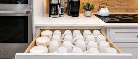 A kitchen with a coffee maker, kettle, and a drawer full of white mugs.