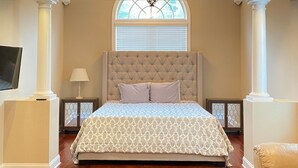 A bedroom with a tufted headboard, a large bed with patterned bedding, wooden flooring, a ceiling fan, and a window with blinds.