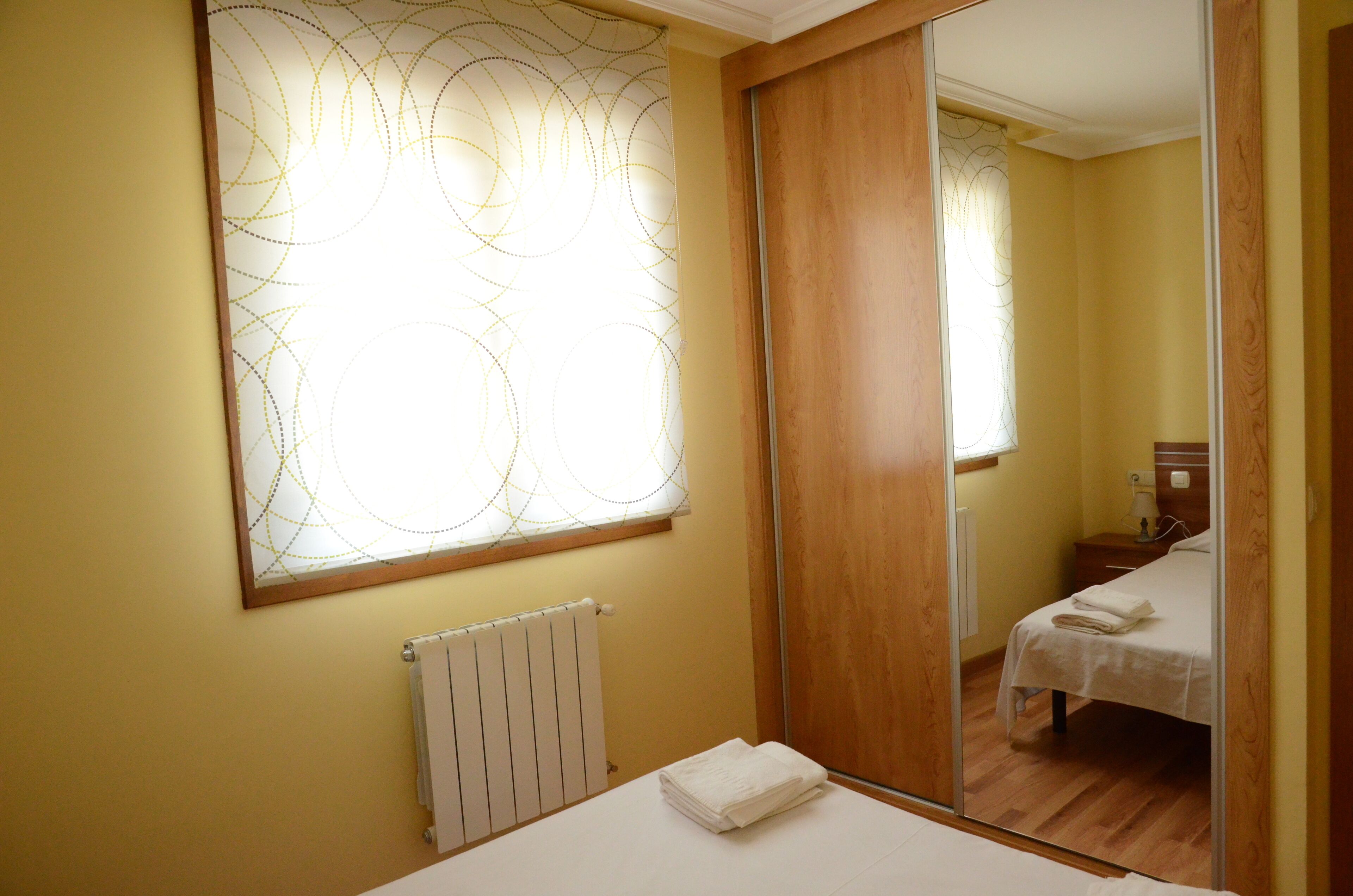 Een slaapkamer met een houten vloer, een witte radiator, een bed met een witte dekbedovertrek en een grote spiegel die de kamer weerkaatst.