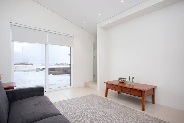 Una sala moderna con un sofá gris, una mesita de madera y un amplio ventanal corredero que da a un paisaje nevado al aire libre.