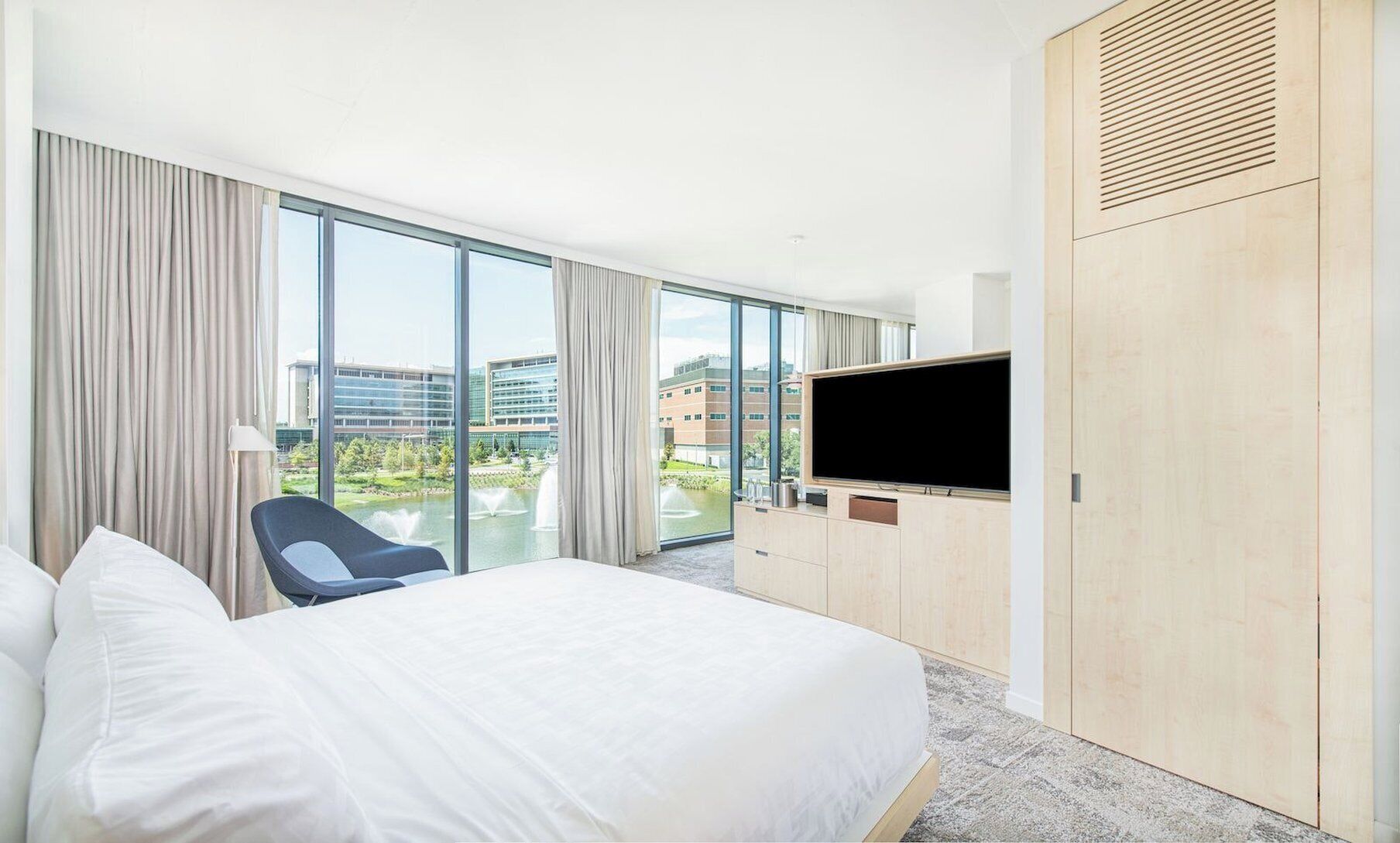 A hotel room with a large bed, a TV, a chair, and a view of a building and water feature.