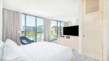 A hotel room with a large bed, a TV, a chair, and a view of a building and water feature.