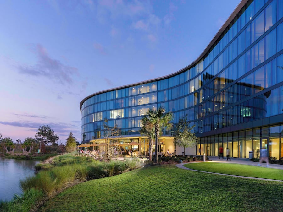 A modern building with a curved glass facade, outdoor seating area, and a grassy courtyard.