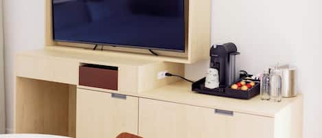 Une pièce comprenant un téléviseur fixé à un meuble en bois, une machine à café, une petite table avec une chaise, et un plan de travail avec une bouilloire et des bouteilles.