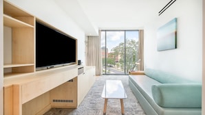Un soggiorno moderno con una televisione a schermo piatto, scaffali in legno, un tavolino, un divano e un'ampia finestra con vista sulla città.