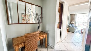 A room with a wooden desk, a mirror, and a vase with branches. There is a door leading to a balcony with a sofa and a view of the outside. - Luana Kai Resort C202 (Kihei)