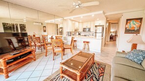 A spacious living area with a television, wooden furniture, and a kitchen in the background. - Luana Kai Resort C202 (Kihei)
