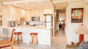 A modern kitchen with a central island, bar stools, and a dining area with a table and chairs. - Luana Kai Resort C202 (Kihei)
