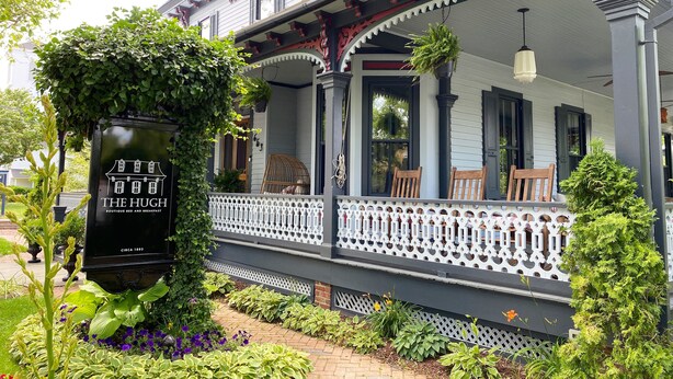 Un edificio histórico con un porche cubierto, un letrero que dice 'The Hugh Boutique Bed and Breakfast' y un jardín bien cuidado con diversas plantas y flores.