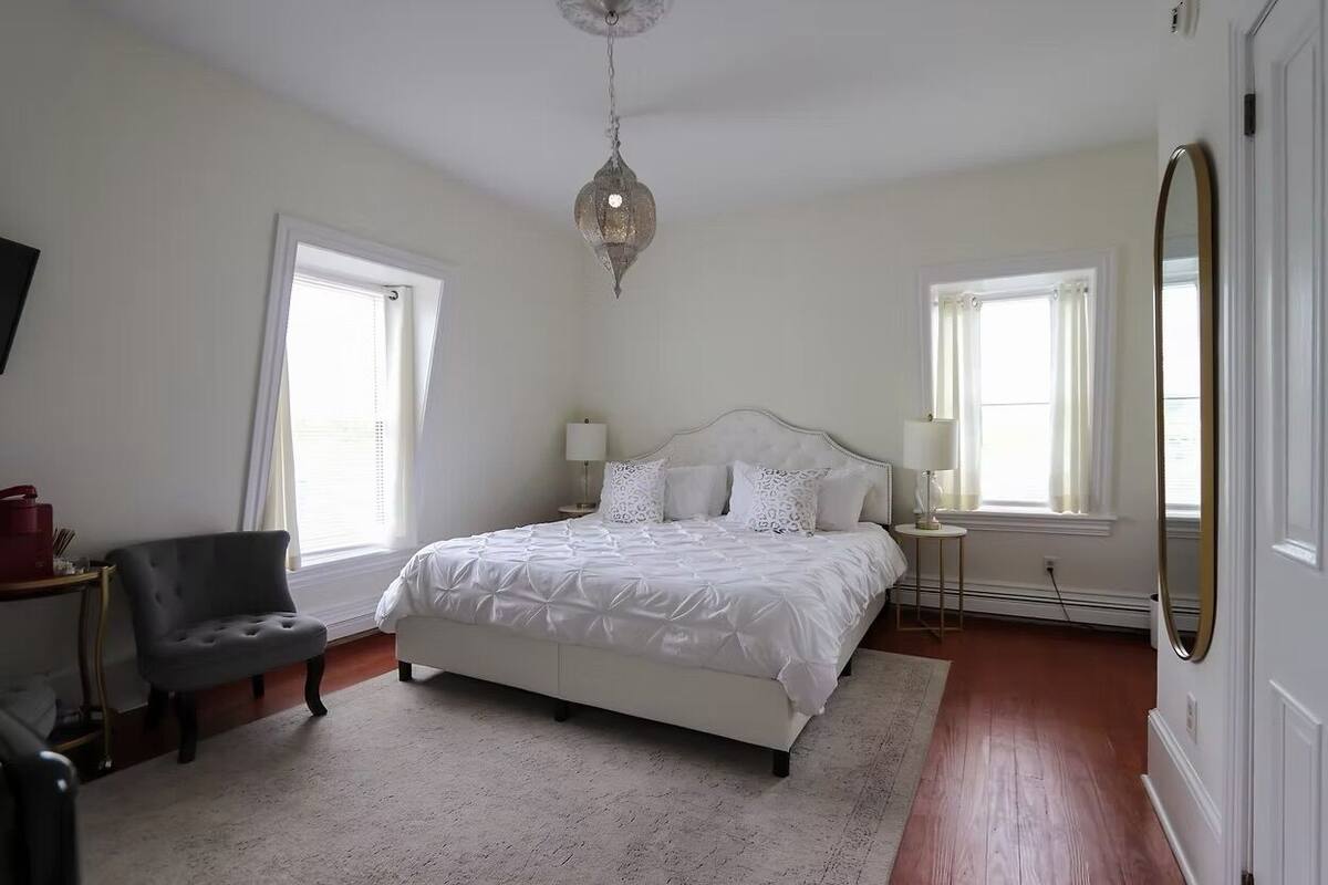 A bedroom with a bed, two bedside tables, a chair, a mirror, and a hanging light fixture.