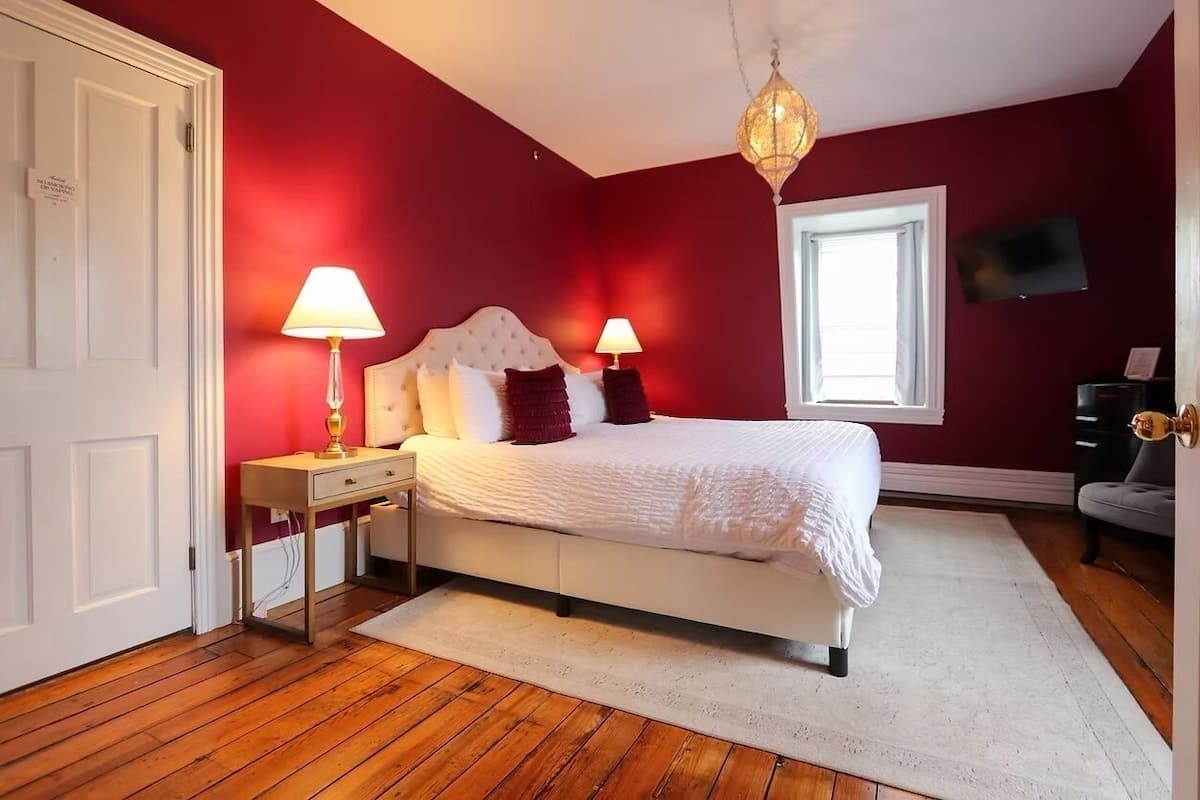 A bedroom with a white bed, two bedside lamps, a red wall, a wooden floor, a white door, and a window with a view.
