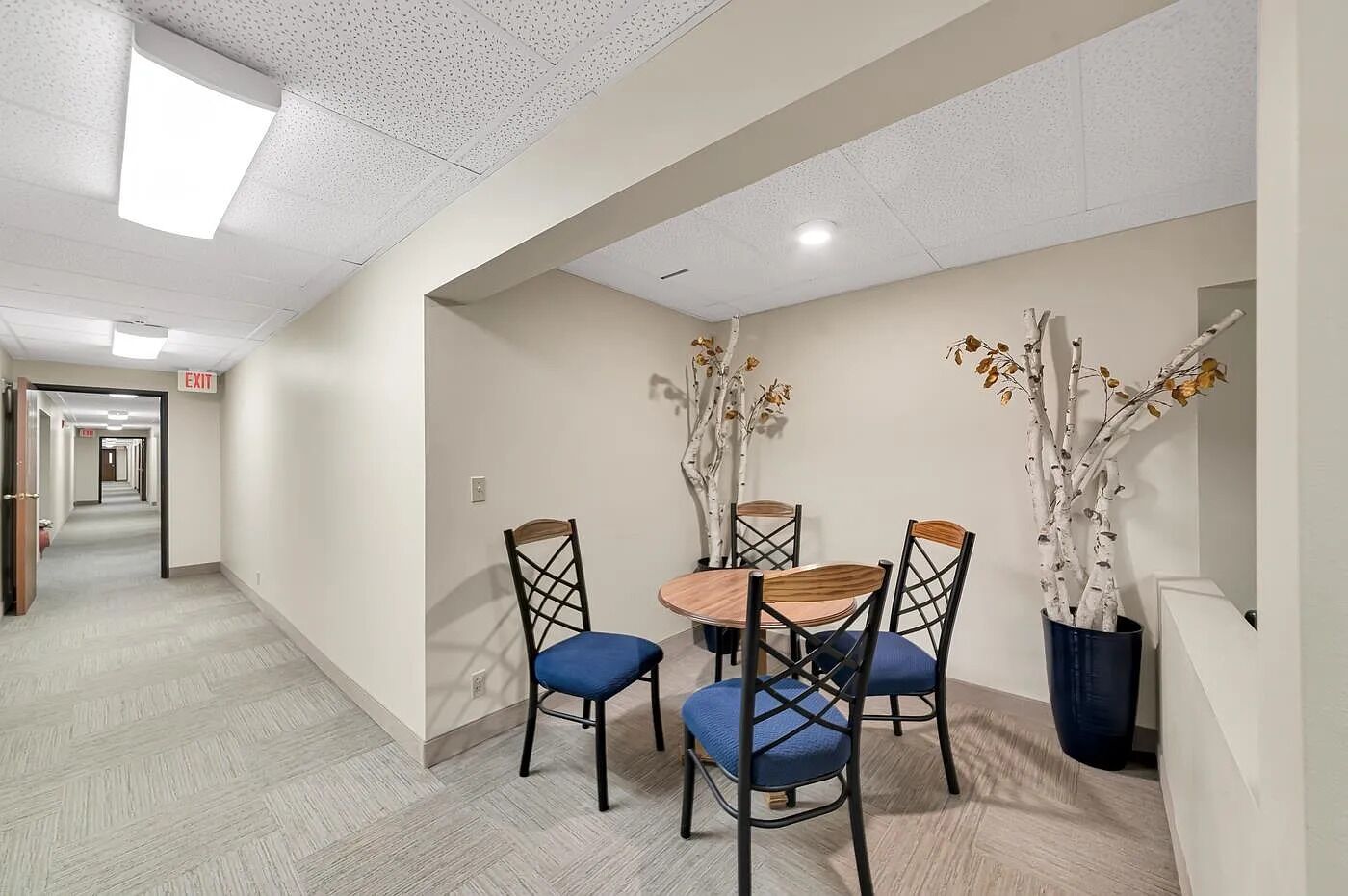 A hotel room with a dining area featuring a round table and four chairs, a potted plant, and a hallway with an exit sign.