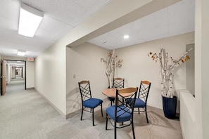 A hotel room with a dining area featuring a round table and four chairs, a potted plant, and a hallway with an exit sign.