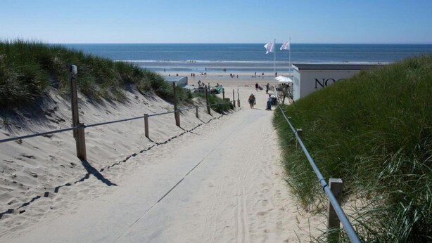Un sentiero sabbioso che conduce a una spiaggia con bandiere e un edificio con la scritta "NOO".