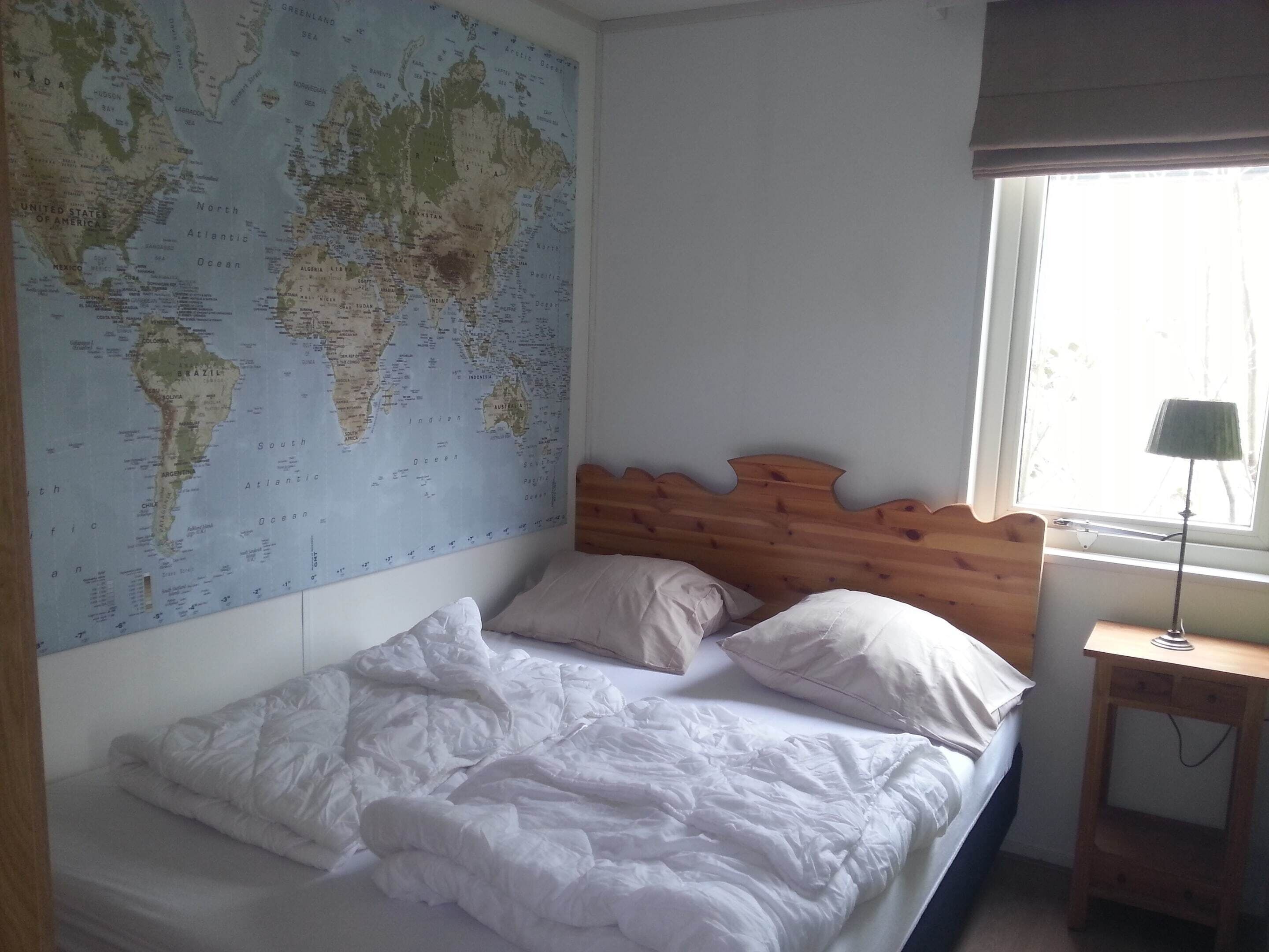 A bedroom with a large world map on the wall, a wooden headboard, a bed with white bedding, and a bedside table with a lamp.