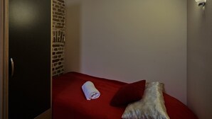 Een klein, keurig opgemaakt bed met een rode sprei, een opgerolde witte handdoek en een decoratief kussen.