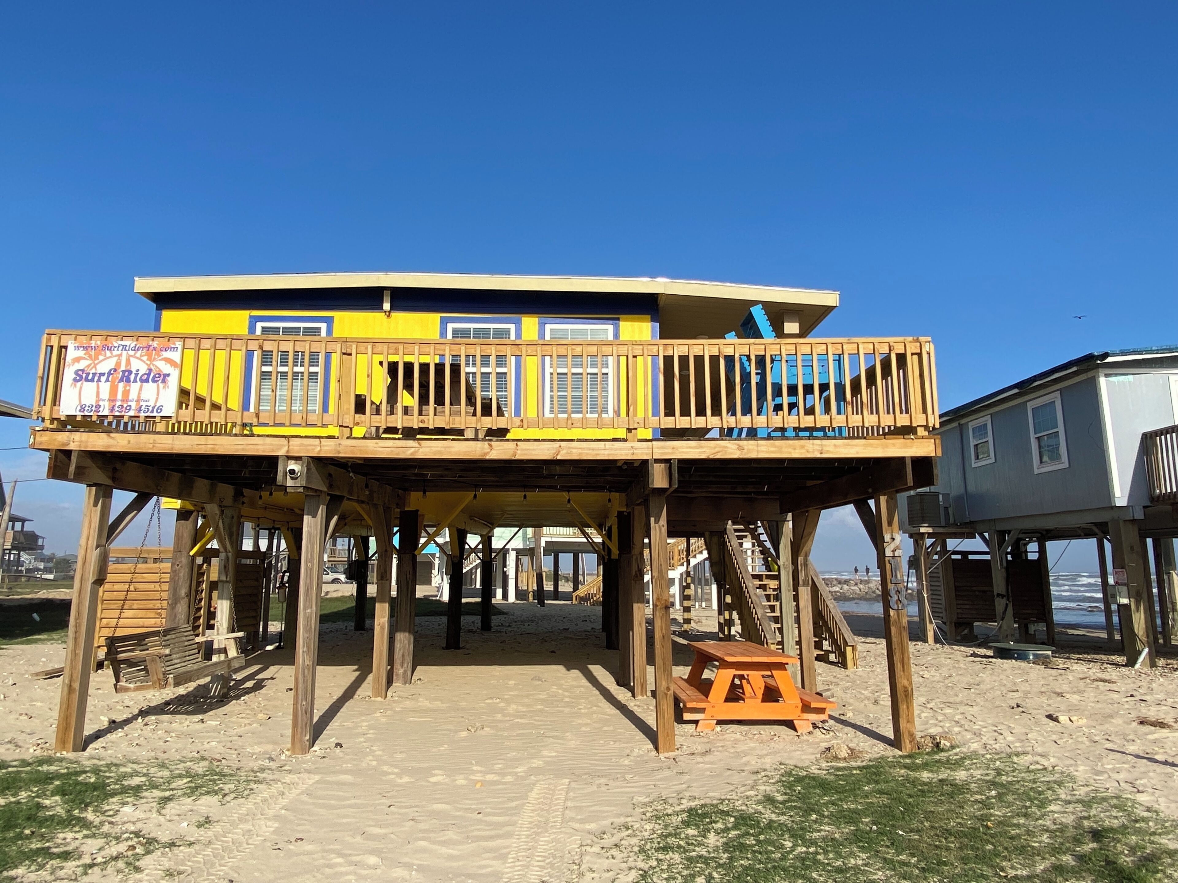 Une structure de terrasse en bois avec un bâtiment bleu et jaune sur pilotis, un banc en bois et une table de pique-nique.
