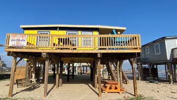 Une structure de terrasse en bois avec un bâtiment bleu et jaune sur pilotis, un banc en bois et une table de pique-nique.