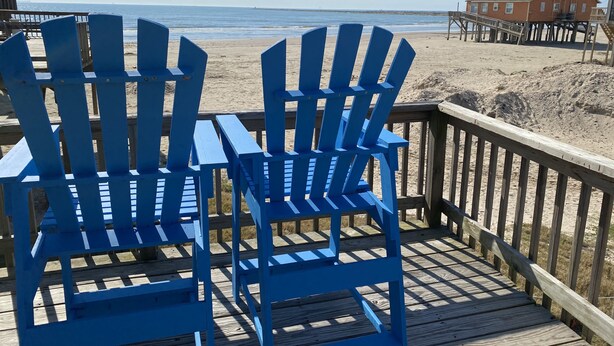 Due poltrone Adirondack blu su un pontile di legno che si affaccia su una spiaggia, con un molo e una casa sullo sfondo.
